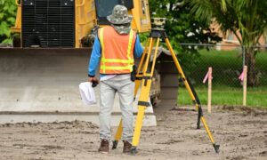 Surveyor conducting a boundary survey on a cleared urban site with safety gear and tripod equipment
