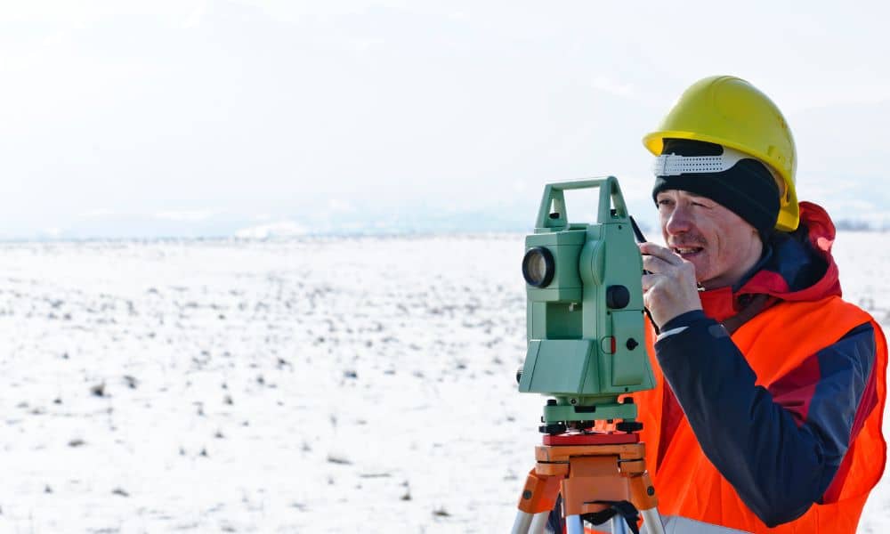 A land surveyor using a theodolite to perform a boundary line survey on a snowy field before the ground freezes